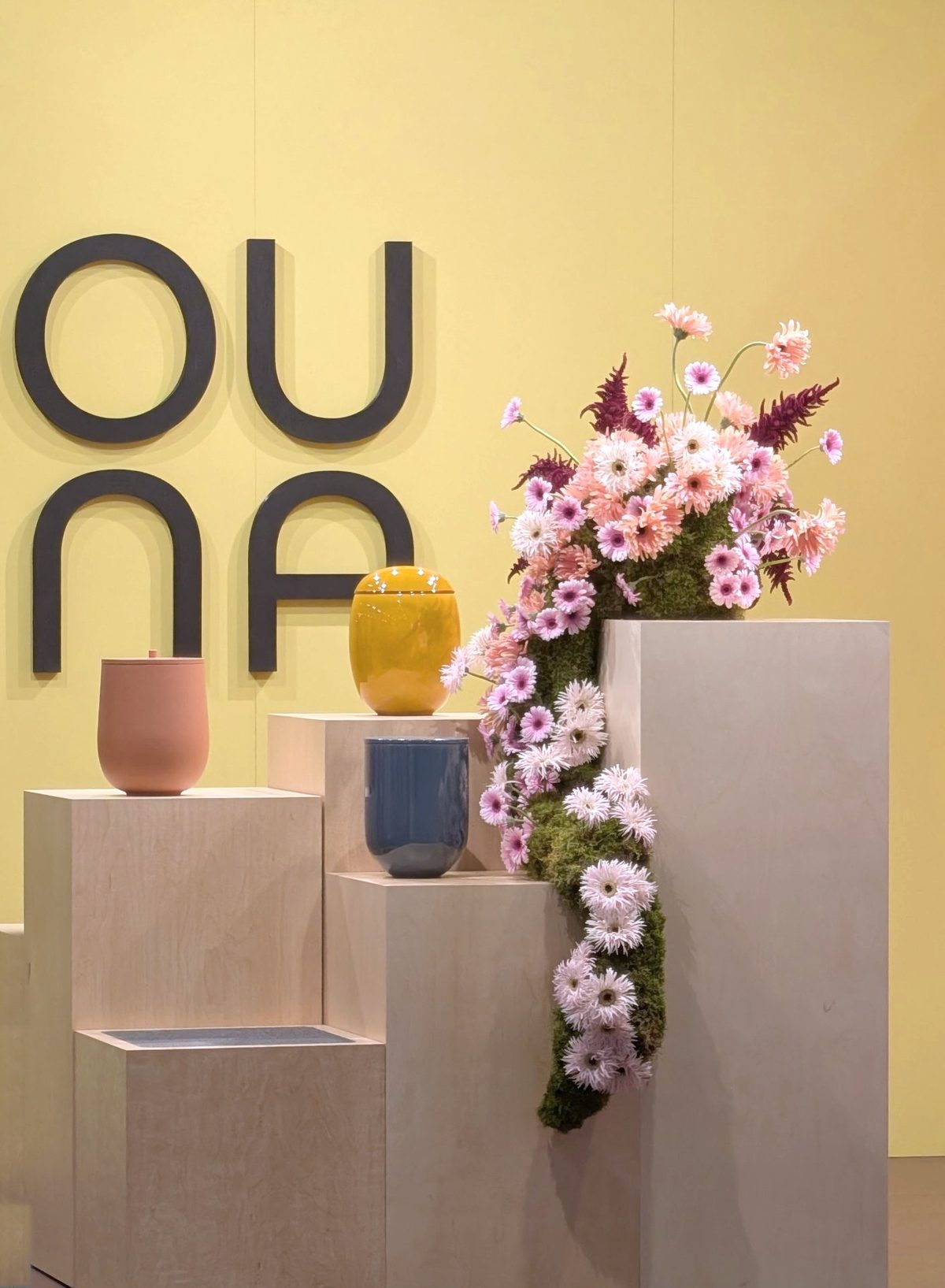 OUNA Urnen auf Holzpodesten vor gelber Wand mit Blumeninstallation